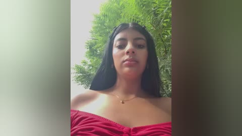 Media: A video of a young woman with long black hair, wearing a red off-shoulder top, standing outdoors against a backdrop of lush greenery.