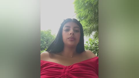 Media: Video of a young woman with medium skin tone and long black hair, wearing a red off-shoulder top, standing in a bright, green outdoor setting.