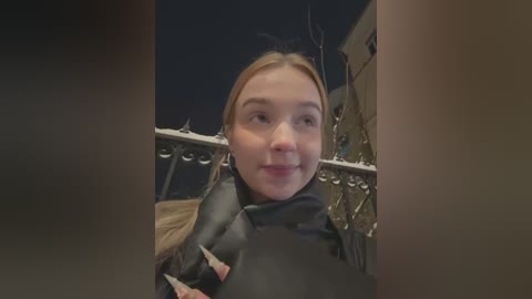 Media: Video of a young Caucasian woman with straight, blonde hair, wearing a black jacket, standing on a balcony at night. Background features ornate metal railings and a dimly lit, beige building.