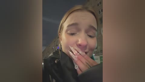 Media: A video of a young woman with fair skin, long blonde hair, and closed eyes, biting her manicured nails while wearing a black leather jacket, set against a blurred urban background.