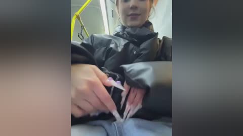 Media: Video of a woman with light skin, light brown hair, and manicured nails, wearing a black puffer jacket, seated on a yellow bus. She is adjusting her clothing, showing a glimpse of her jeans.