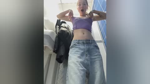 Media: Video of a tall, slim woman with light skin and blonde hair, wearing a purple crop top and light blue jeans, lifting her shirt, standing in a small bathroom with a black jacket hanging on a hook.