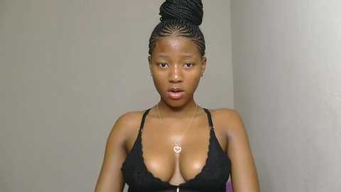 Video of a dark-skinned woman with an athletic physique, wearing a black lace bra, her hair styled in a high bun. She has a small, silver necklace. Plain beige wall background.