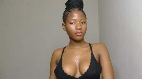 Video of a young Black woman with medium-dark skin and a slender build, wearing a black lace bralette, her hair styled in a high bun. She stands against a plain, light-colored wall.