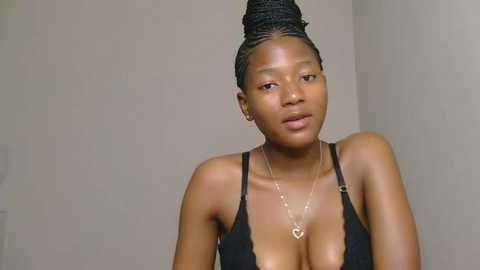 Media: Video of a young Black woman with a medium complexion and braided hair in a high bun, wearing a black spaghetti-strap top and a gold heart necklace.