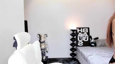 Media: Video of a minimalist bedroom with a white leather chair, a bear statue, and a black cube lamp on a gray bed.