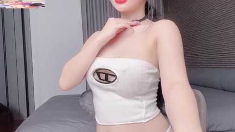 Video of a fair-skinned woman with red lipstick, wearing a white, strapless crop top with a large black \"T\" emblem, in a modern bedroom with gray bedspread and blinds.