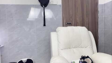 Media: Video of a modern bathroom with light gray tiled walls, a white leather recliner, wooden door, black hanging robe hook, and a black shoe on the floor.