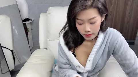 Media: Video of a young Asian woman with fair skin, wearing a light gray robe, revealing a black bra underneath, sitting on a white leather chair in a modern, minimalistic room with gray walls and a wooden door.