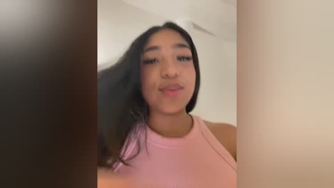 Media: A video of a young Latina woman with long black hair, wearing a pink tank top, standing in front of beige walls, with blurred vertical objects on either side.