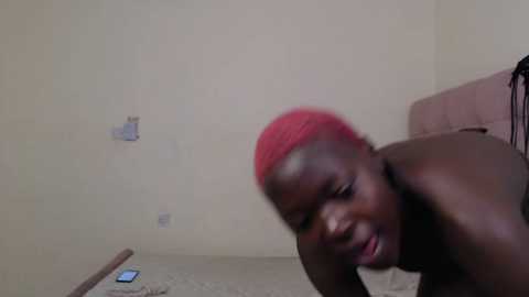 Media: Video of a dark-skinned man with red hair, topless, lying on a beige carpet in a sparsely furnished room with cream walls and a beige couch.