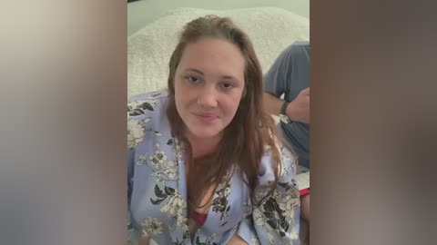 Media: Video of a fair-skinned woman with long brown hair, wearing a floral-patterned blue robe, sitting on a white carpeted floor. She has a gentle smile and her arms are crossed.