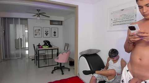 Media: Video of a modern, minimalist apartment with a man taking a selfie in the foreground. In the background, a woman sits on a chair, wearing a white tank top and shorts, next to a black office chair.