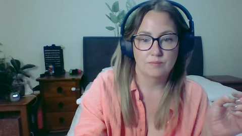 Media: Video of a woman with straight, light brown hair, wearing black-framed glasses and a pink shirt, sitting in a bedroom with a wooden dresser, potted plants, and a bed in the background.