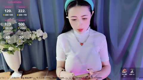 Media: Video of a young East Asian woman with long black hair, wearing a white top, teal headband, and necklace. She sits in front of a blue curtain with a vase of white flowers behind her.