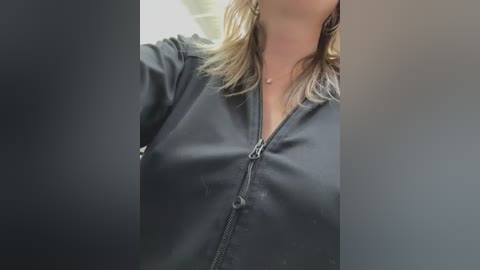 Media: Video of a woman with light skin, shoulder-length blonde hair, wearing a black zip-up jacket, with a pendant necklace visible.