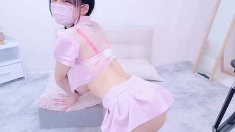 Media: Video of an East Asian woman in a pastel pink nurse cosplay, wearing a face mask, kneeling on a bed in a minimalist, light-colored room.