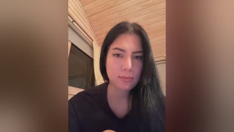 Media: A video of a young woman with long, dark hair, fair skin, and a neutral expression, wearing a dark shirt, standing in a room with wooden paneling and a window with a closed blind.