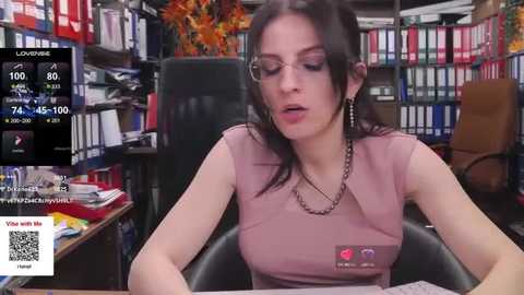 Media: Video of a young, pale-skinned woman with long black hair and glasses, wearing a pink top and necklace, sitting at a cluttered desk in a library-like room with bookshelves and a potted plant.