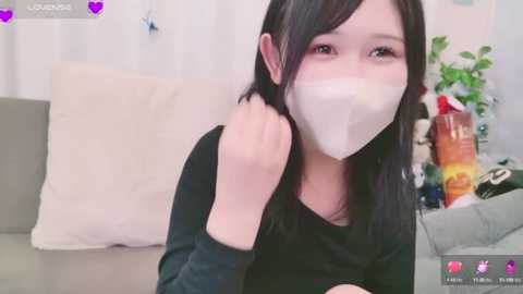 Media: Video of an East Asian woman with light skin, black hair, wearing a white face mask, black top, and smiling, sitting on a beige couch. Background includes a green plant, a bottle, and white curtains.