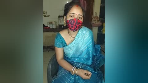 Media: Video of a woman in traditional Indian attire, wearing a blue sari with a red mask, sitting in a dimly-lit, cluttered kitchen.