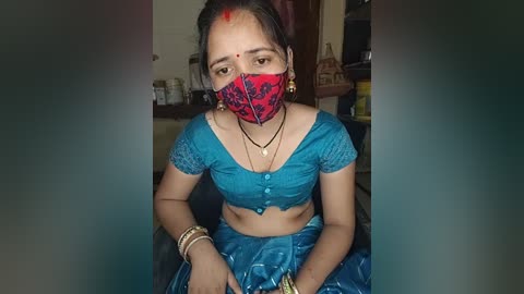 Media: Video of a young woman with medium brown skin, wearing a blue saree, red face mask, and red bindi, sitting indoors with traditional Indian decor in the background.