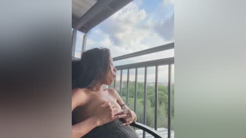 Video of a topless woman with long black hair, sitting on a balcony with a green landscape and blue sky background.