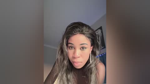 Media: Video of a Black woman with long, wavy black hair and light brown skin, wearing a black top and large silver hoop earrings, looking directly at the camera in a dimly lit room.