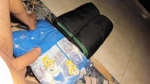 Media: A video shows a man's legs in black socks and blue shorts, sitting on a tiled floor. He's holding a blue T-shirt with cartoon characters. The background features a patterned rug and a black bag.