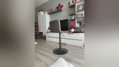 Media: Video of a modern living room with a gray sofa, white TV stand, gray shelves, and a Christmas tree on the left. The floor is light wood, and the background is gray.