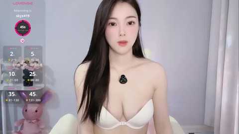 Media: A young Asian woman with long black hair, fair skin, and a small bust, wearing a white bra and necklace, sits in a room with a pink bunny and a white shelf.