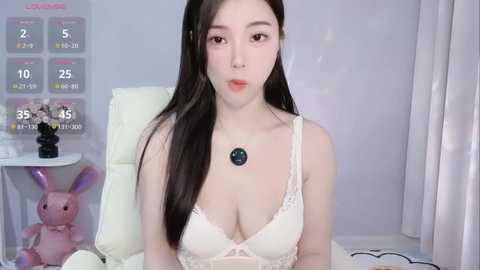 Media: Video of an East Asian woman with long black hair, wearing a white lace bra, sitting on a white bed, with a calendar and plush toys in the background.