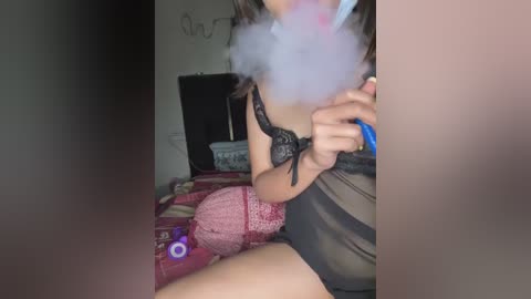 Media: A video of a woman with light skin, long brown hair, and wearing a sheer black lace bra, blowing smoke from a vape. She sits on a bed with a pink blanket and a purple toy. The background shows a dark wall with a white shelf.