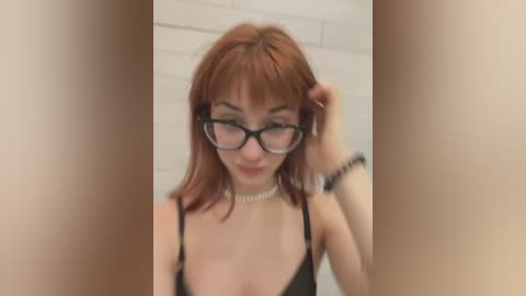 Media: Video of a young woman with fair skin, shoulder-length auburn hair, and wearing black-framed glasses, adjusting her hair. She has a slender build and is dressed in a black spaghetti-strap top. Background shows a white brick wall.