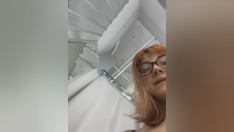 Video of a young Caucasian woman with light skin, long red hair, and black-rimmed glasses, wearing a white top, standing in a modern, minimalist room with white walls and a spiral staircase.