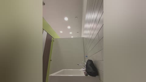 Video of a narrow, modern bathroom with white tiles, a white sink, and a lime green accent wall. A black soap dispenser is mounted on the right wall.