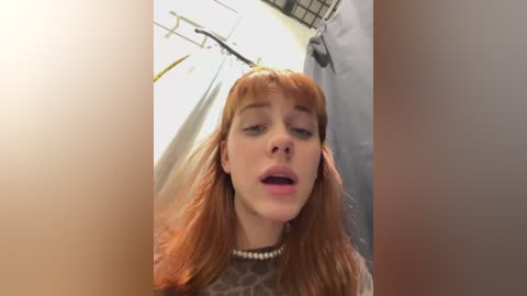 Video of a fair-skinned, red-haired woman with blunt bangs and a pearl necklace, mouth slightly open, in a bathroom with shower curtain and clothes hanging in the background.