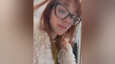 Video of a young woman with fair skin, red hair, and large black-rimmed glasses, wearing a fluffy white sweater, smiling slightly, with a blurred background.