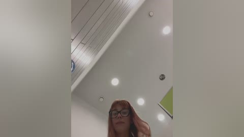 A video of a young woman with long, straight red hair and glasses, standing in a modern, minimalist room with white walls, recessed lights, and a green wall in the background.