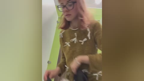 Media: Video of a pale-skinned, pink-haired woman with glasses wearing a brown, animal-print sweater, standing in a green hallway, blurred background.