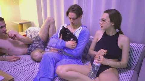 Media: Video of three people on a striped couch: a shirtless man in plaid shorts, a man in blue pajamas holding a black cat, and a woman in glasses and a black swimsuit.