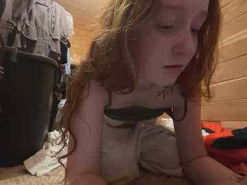 Media: Video of a young, fair-skinned, red-haired woman with long hair, wearing a black bra and beige pants, crawling on a carpeted floor in a cluttered room with wooden paneling, laundry, and a black trash can.