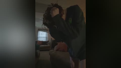 A dimly lit video of a person with curly hair bending over, wearing a dark hoodie, standing in a living room with a couch and window.