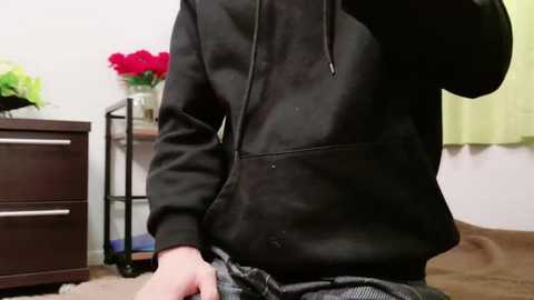 Media: Video of a person in a black hoodie, slightly lifting their shirt to reveal a gray plaid shirt underneath, sitting on a couch with a vase of red roses on a dark wooden dresser in the background.