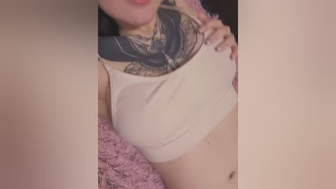 Media: Video of a light-skinned woman with long black hair, wearing a beige tank top revealing a detailed black butterfly tattoo on her chest, and a pink fuzzy blanket in the background.