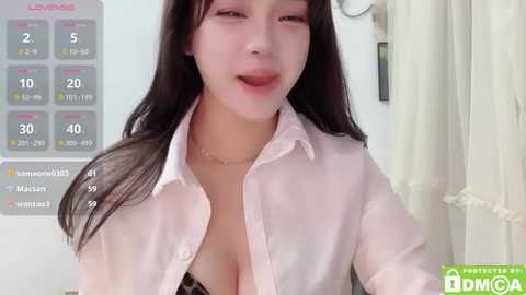 Media: A video of a young Asian woman with long black hair, wearing a semi-transparent pink blouse, revealing cleavage, in a modern, well-lit room.