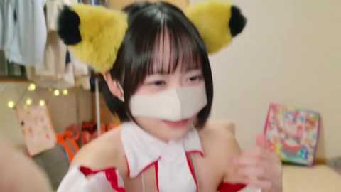 Video of an Asian woman with short black hair wearing a white mask, red kimono, and yellow fox ears, giving a thumbs-up gesture. Background shows a cluttered room with hanging clothes and festive decorations.