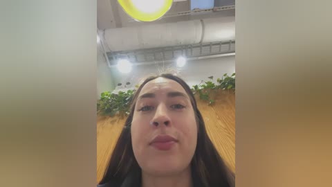 Video of a young woman with long brown hair, medium skin tone, and neutral expression, standing in a modern indoor setting with a wooden table, green plants, and overhead lighting.