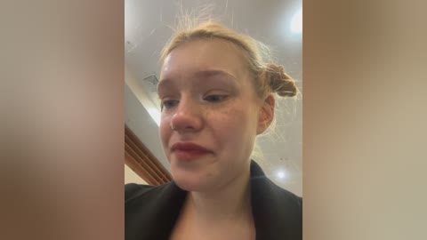 Media: Video of a young Caucasian woman with light skin, blonde hair in a bun, wearing a black jacket. She has a neutral expression. The background shows an indoor setting with wooden beams and white ceiling.