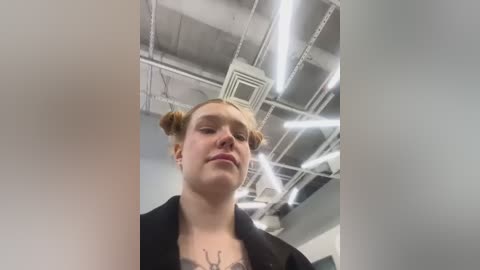 Media: Video of a young woman with fair skin and blonde hair styled in two buns, wearing a black coat, standing in a modern, industrial room with exposed ceiling ducts and fluorescent lights.